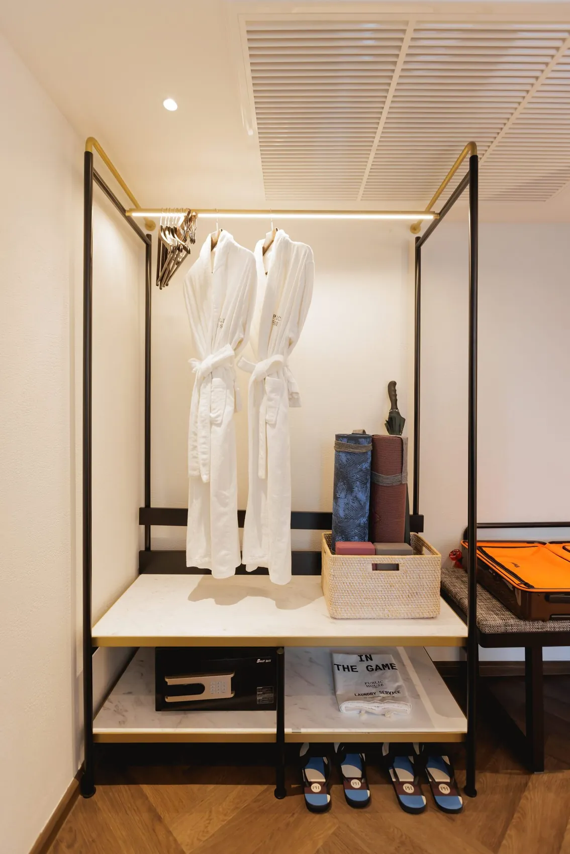Open wardrobe in the Junior Suite half of the Two-Bedroom Suite with bathrobes on a brass rail, the in-room safe, slippers and a luggage bench