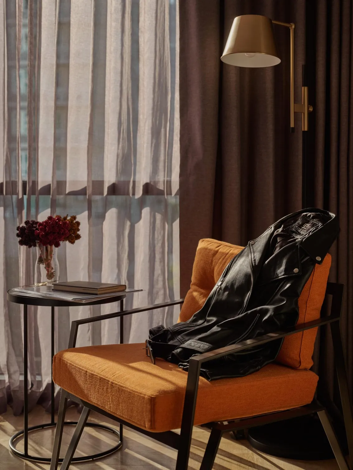 Orange armchair beside a curtained window, with a brass reading lamp and a vase of flowers
