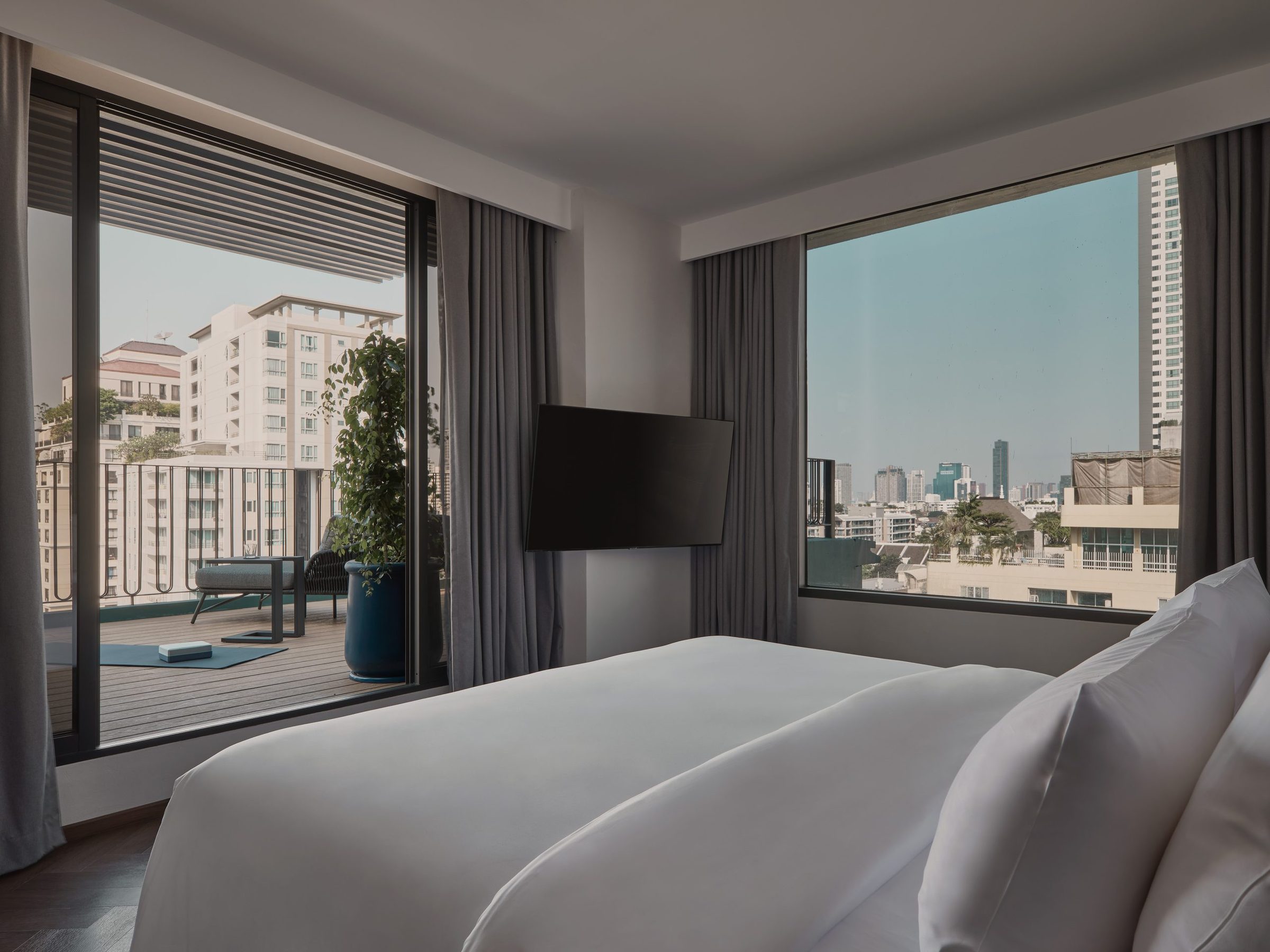 A bed at Public House Bangkok looking out through full-height glass to a private terrace and the Bangkok rooftops