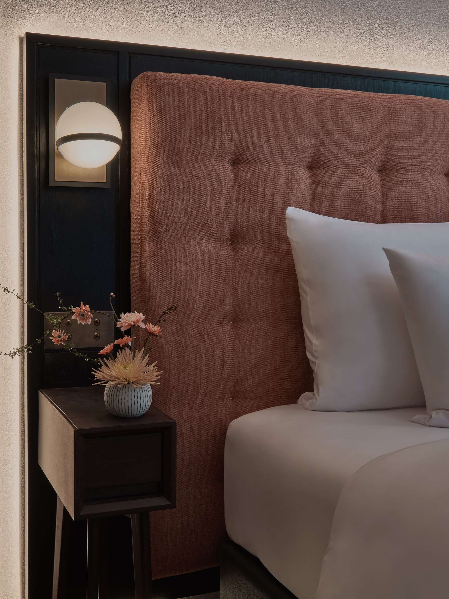 Bed with a terracotta buttoned headboard, a globe wall light and fresh flowers on the nightstand