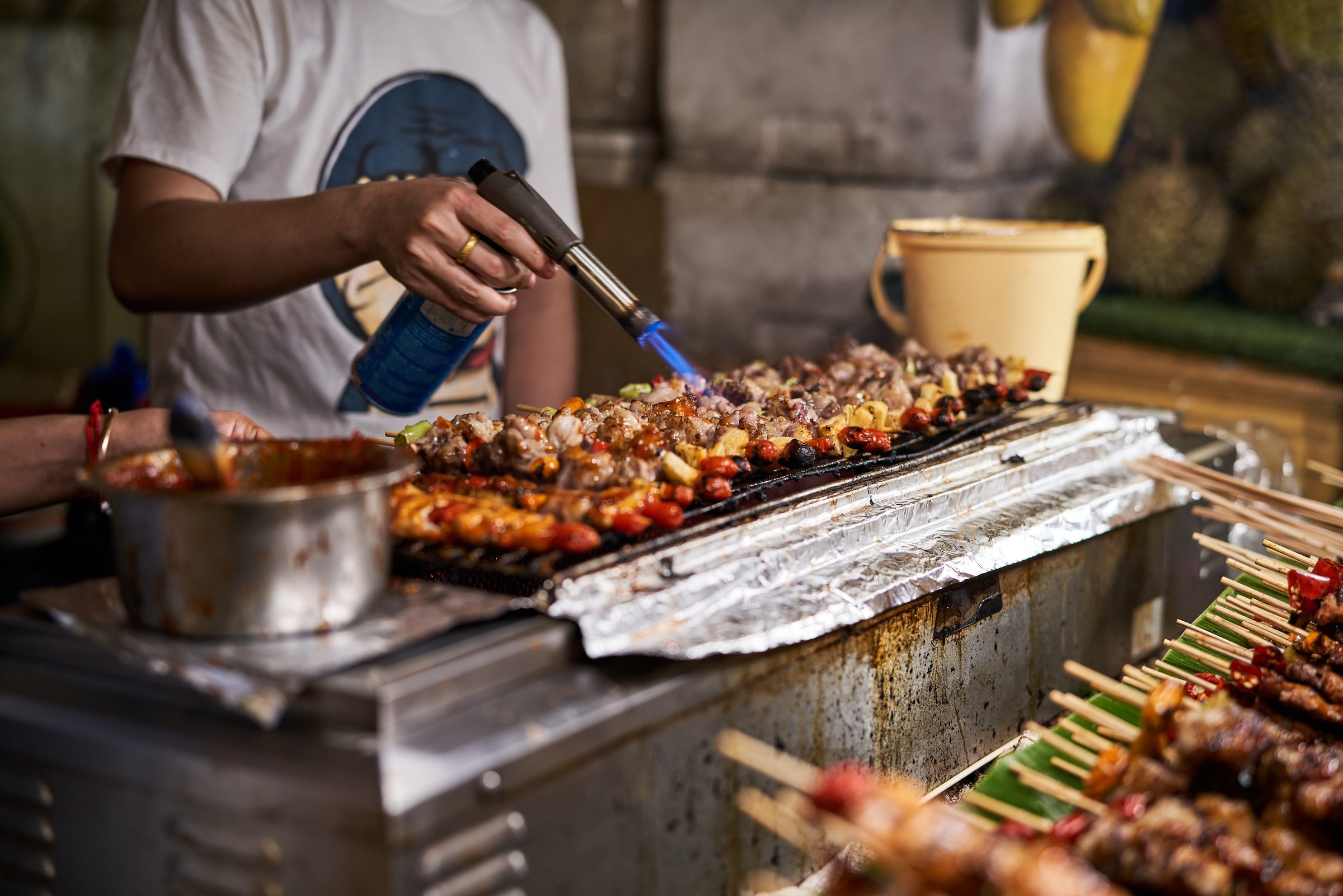 A vendor finishing grilled skewers with a torch at a Bangkok street stall