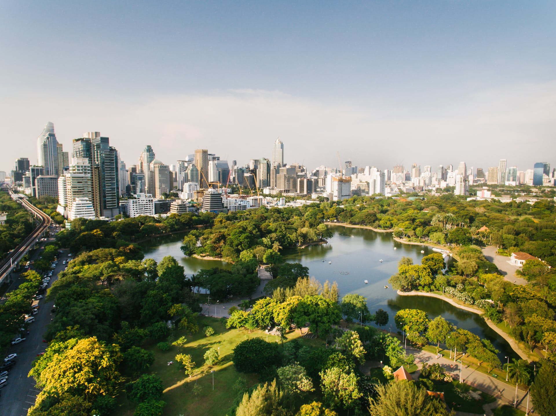 An aerial view of a Bangkok park, its lake ringed by trees with towers behind