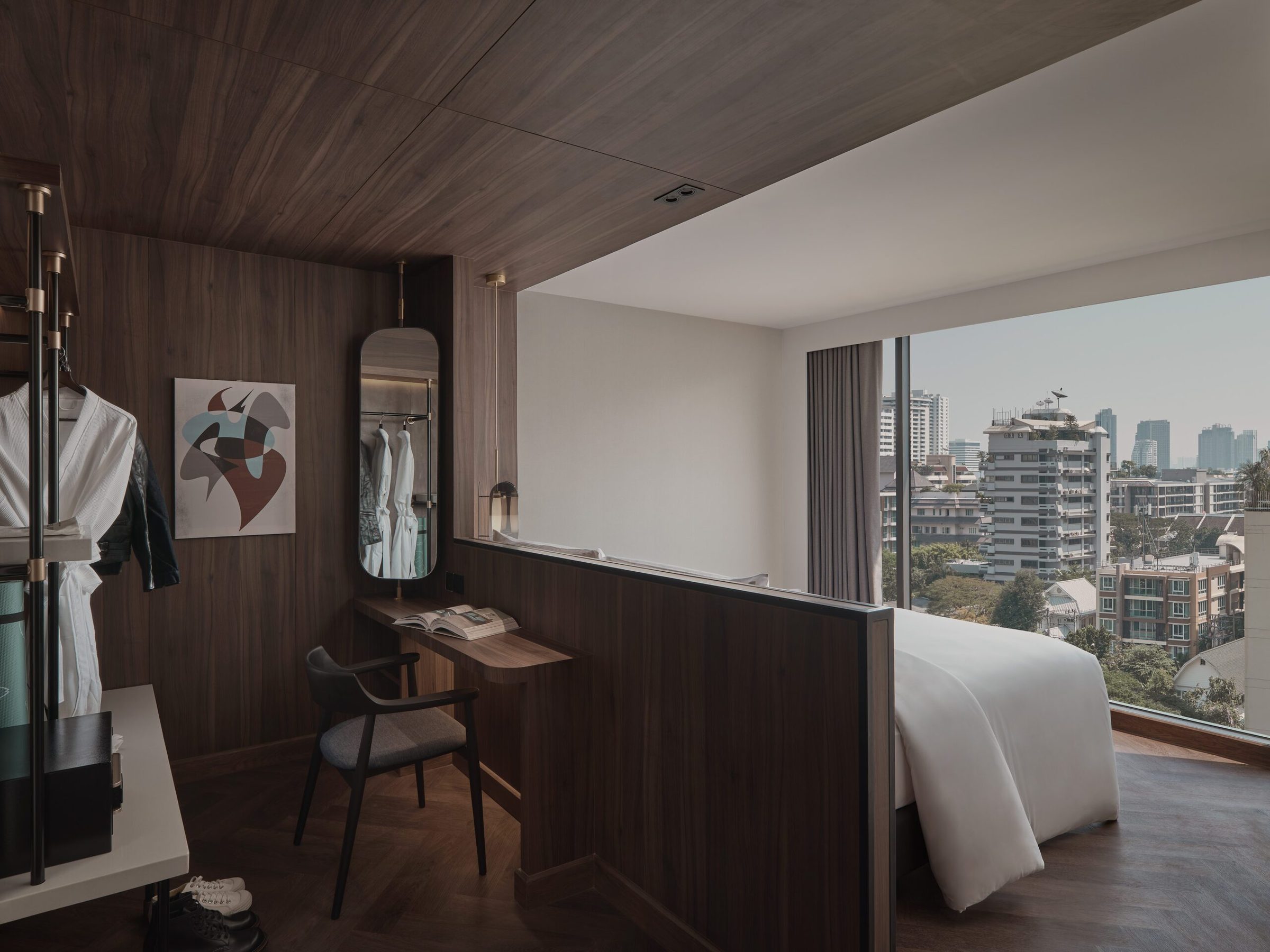 Executive Suite at Public House Bangkok: a walnut-panelled dressing area with a writing desk and mirror, opening past the bed to a full-height city window