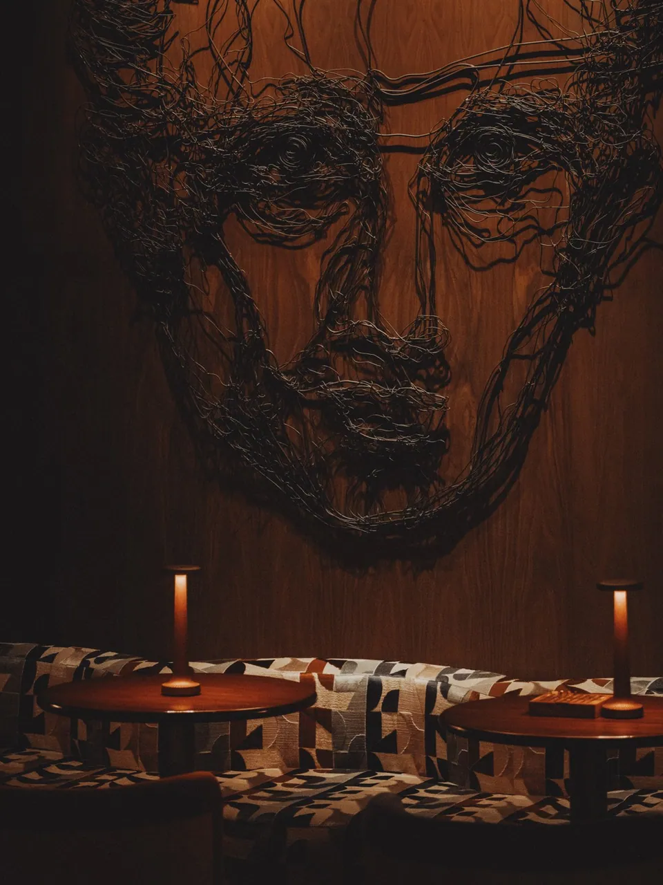 A wire portrait sculpture on the timber wall at OPEN BAR, above candle-lit round tables and orange stools