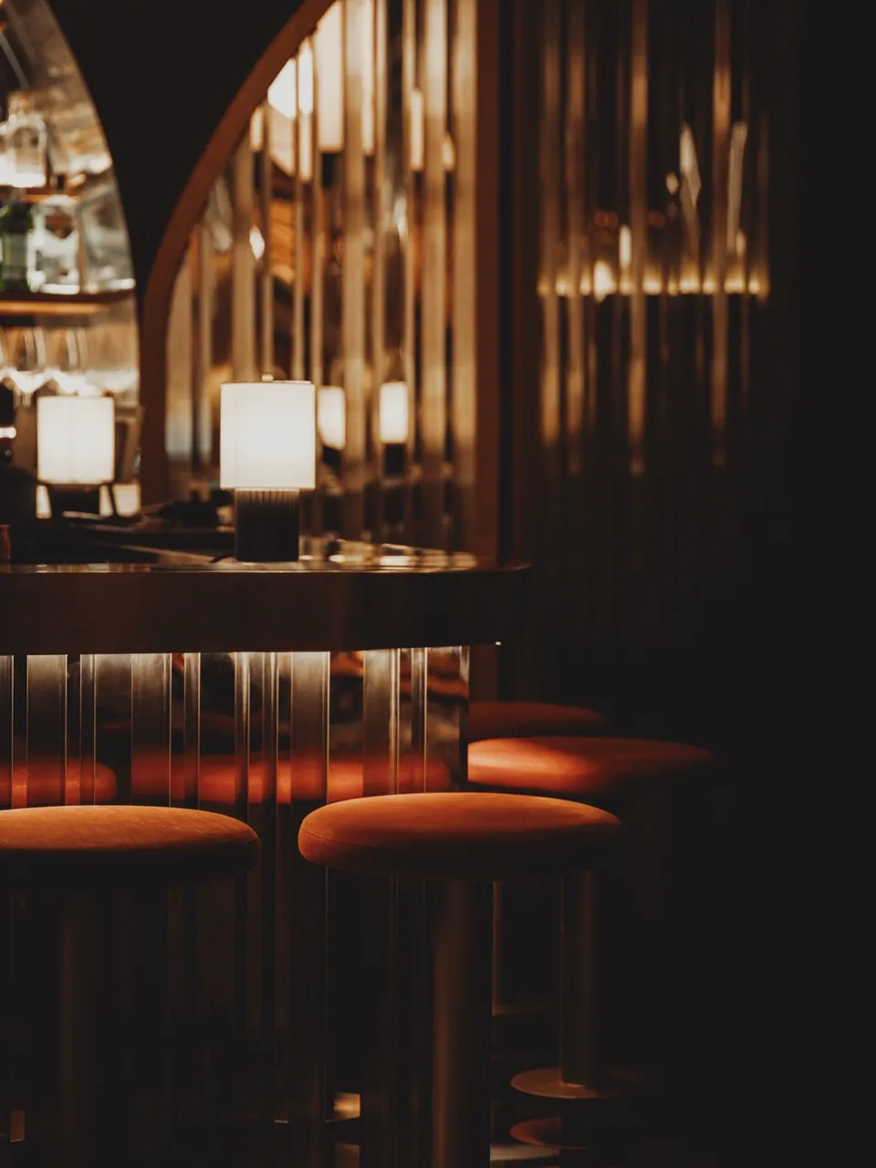 A corner of OPEN BAR, arched shelving lined with bottles and lamps above an orange banquette