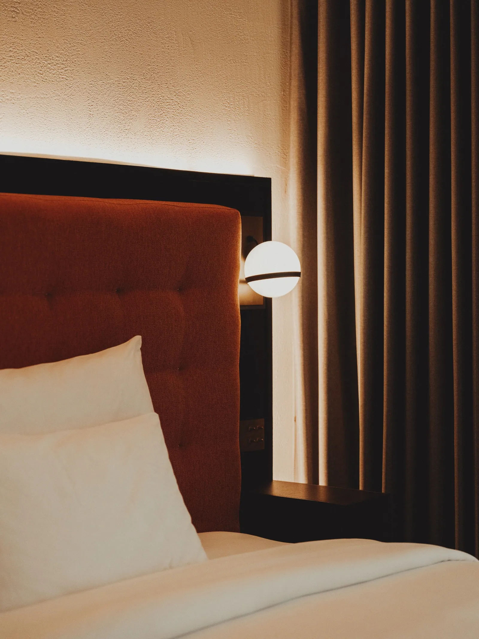 A rust upholstered headboard, a linen pillow and a globe wall light in a guest room at Public House Bangkok