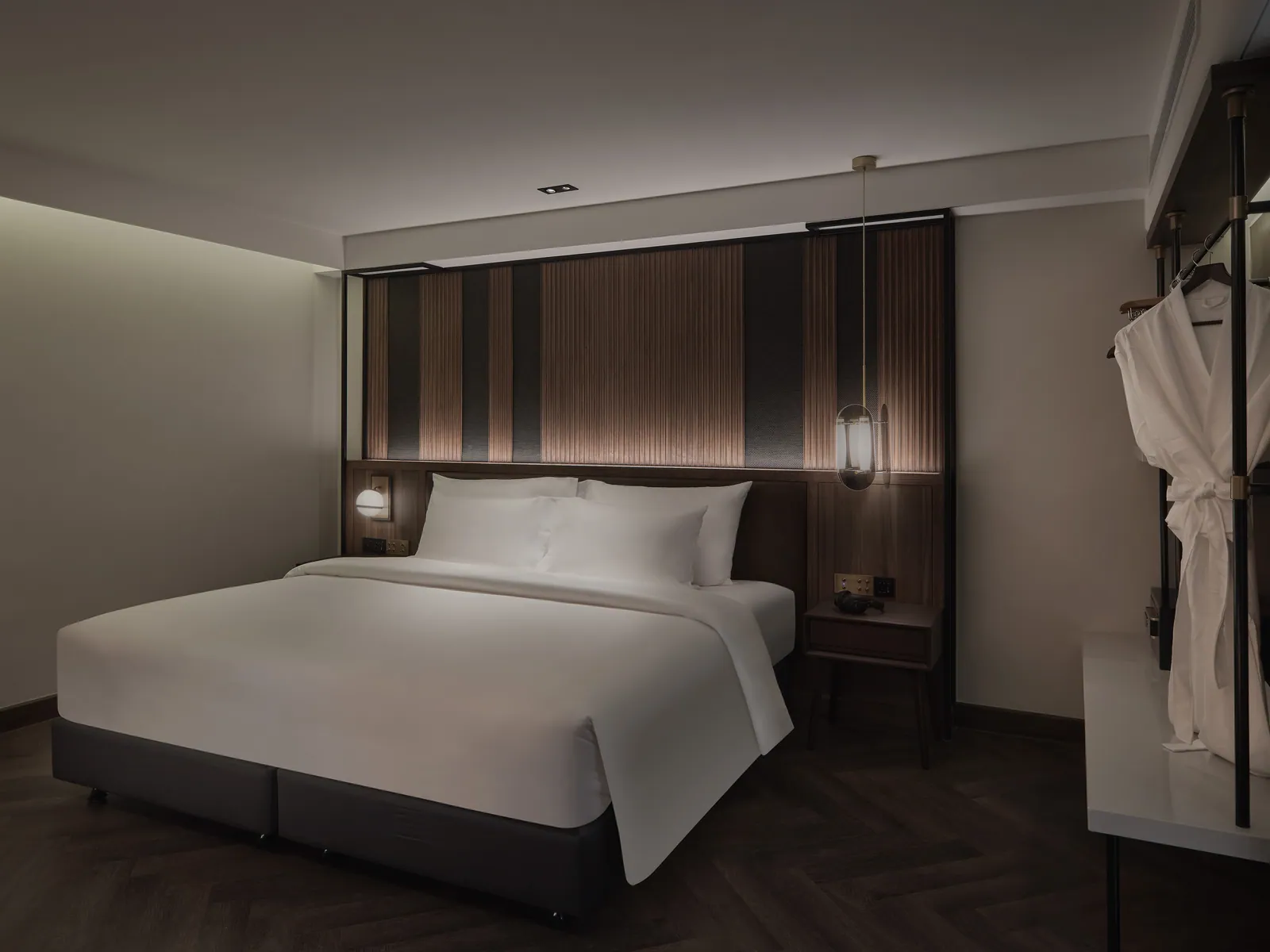 A guest room at Public House Bangkok with a slatted teak headboard, a bedside pendant and a robe hanging by the wardrobe