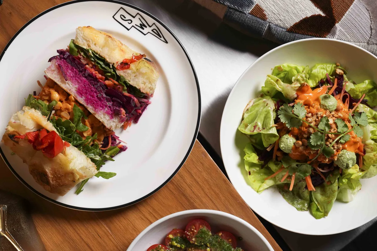 Plates shared across a walnut table at SWD: a layered focaccia sandwich on an SWD plate, a green salad and burrata with tomatoes