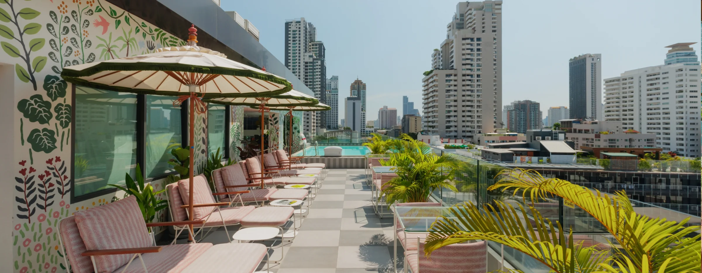 The rooftop at THE MOOON, paper parasols over striped sun loungers running down to the swimming pool with the Sukhumvit skyline beyond