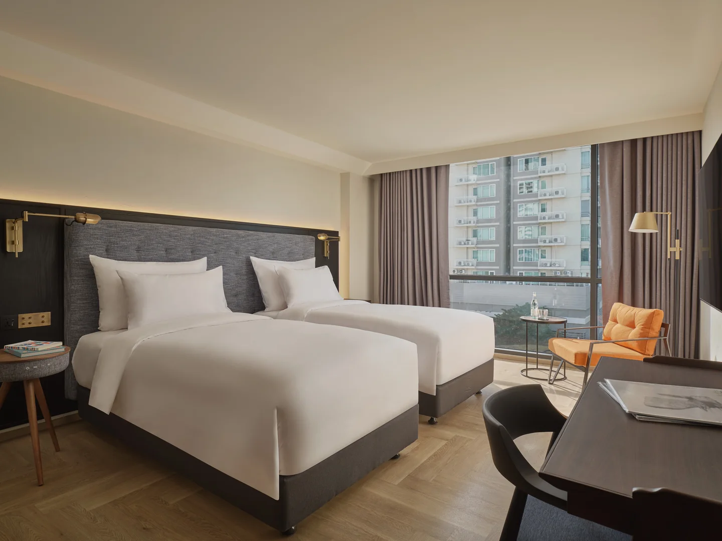 Twin beds, an orange armchair and a Sukhumvit city view through the window of the 28 sqm Uptown Room at Public House Bangkok