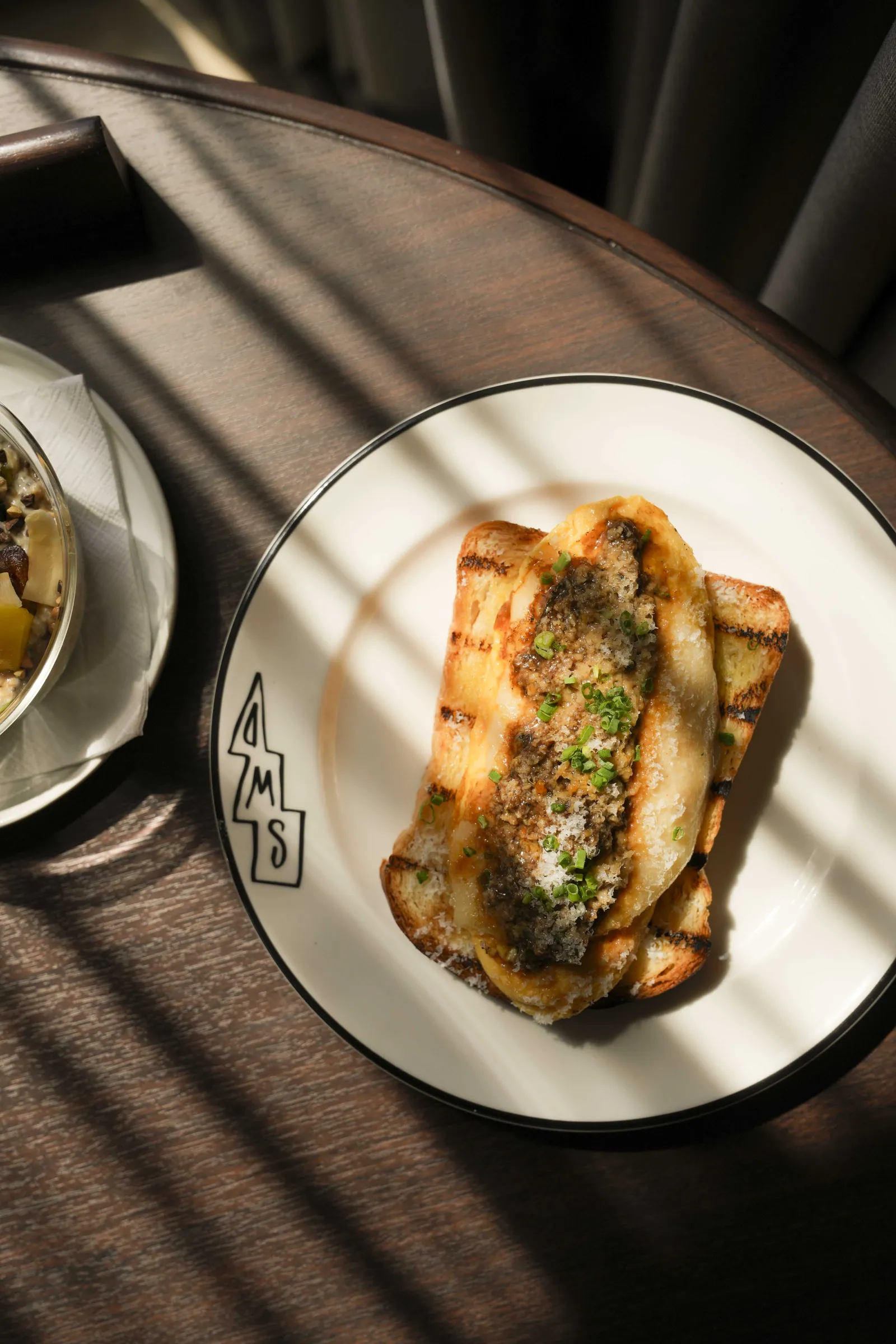 Toasted sourdough under a herbed topping on an SWD plate, on a dark wood table striped with morning sun