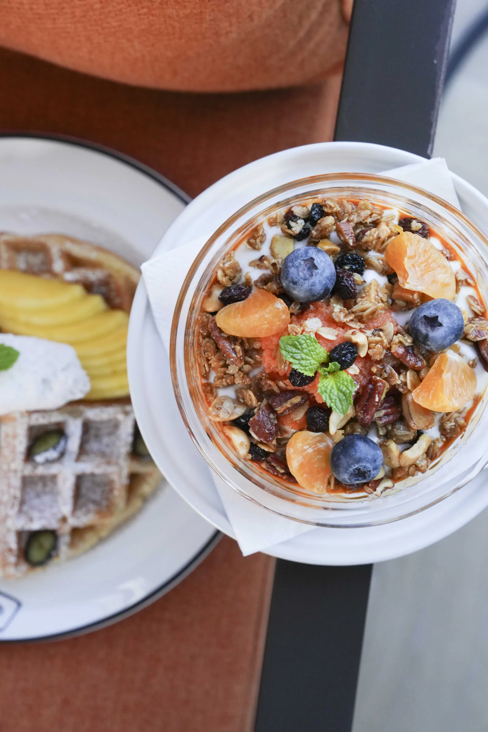 A glass of granola with blueberries, mandarin and mint at SWD, beside a waffle plate and sliced fruit on marble