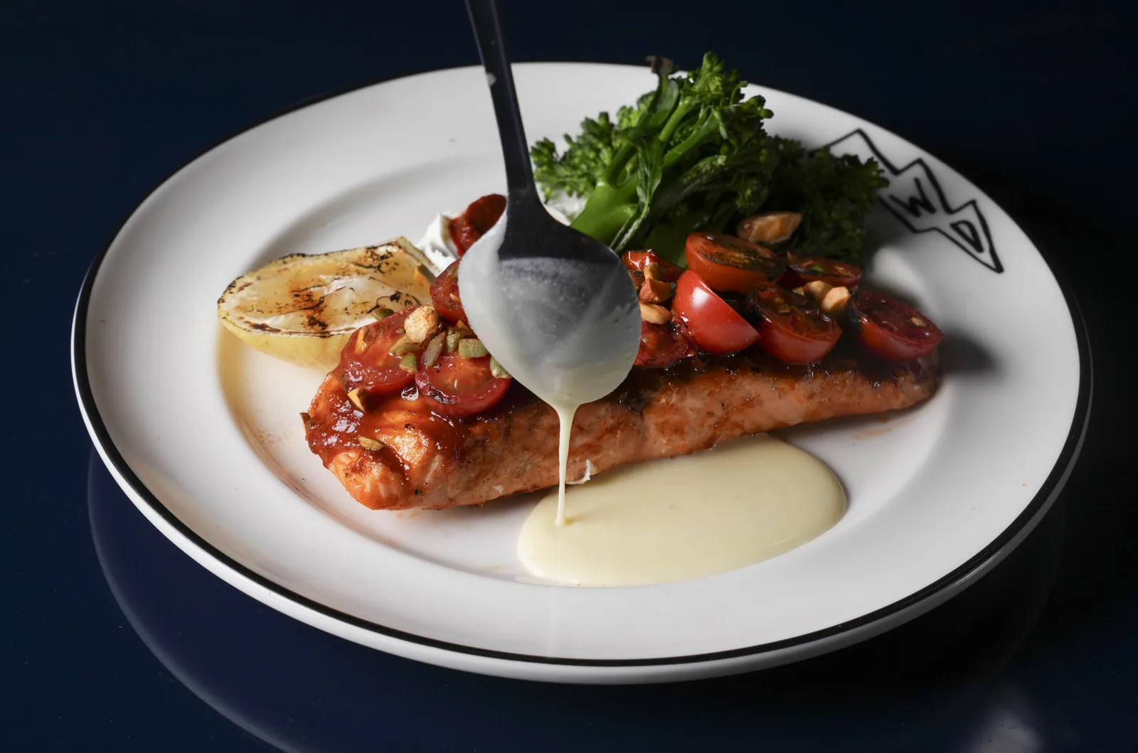 A grilled salmon fillet at SWD with tomatoes, broccolini and a charred lemon half, sauce being spooned over