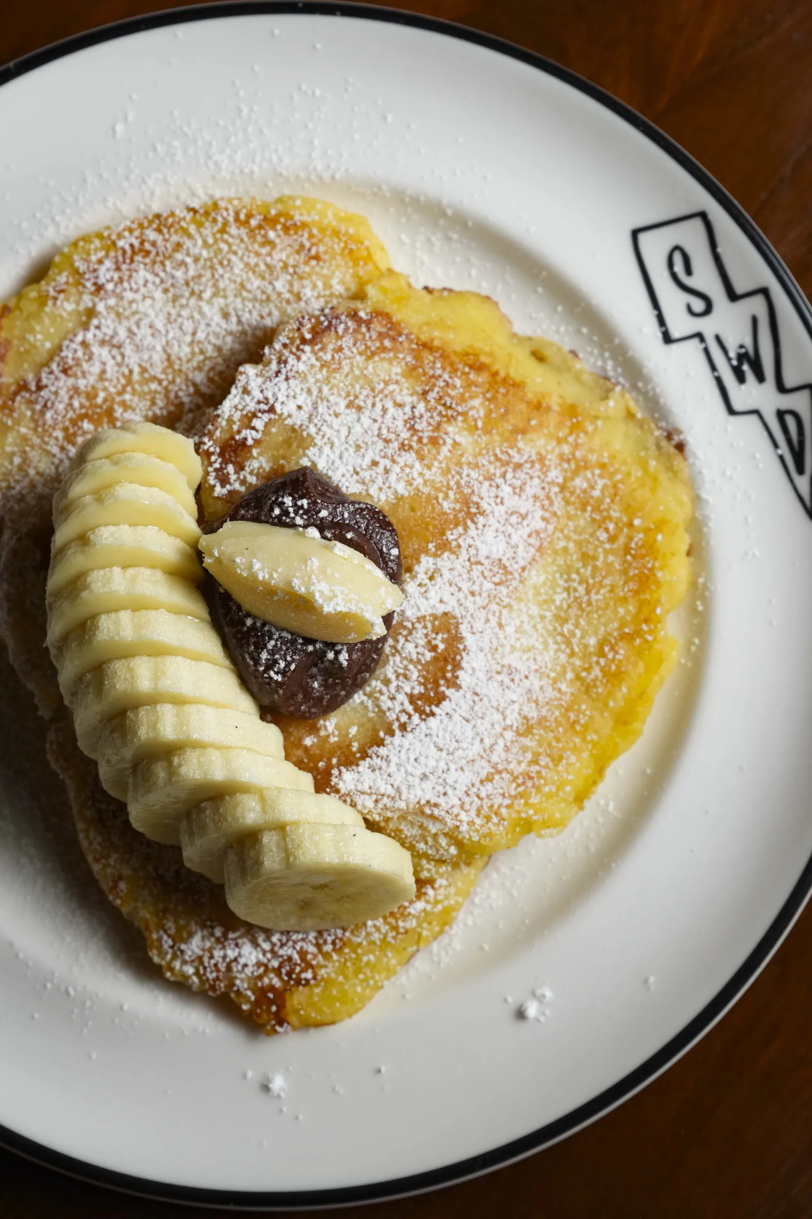 A ricotta pancake at SWD with sliced banana, quenelles of custard and icing sugar, on a branded SWD plate